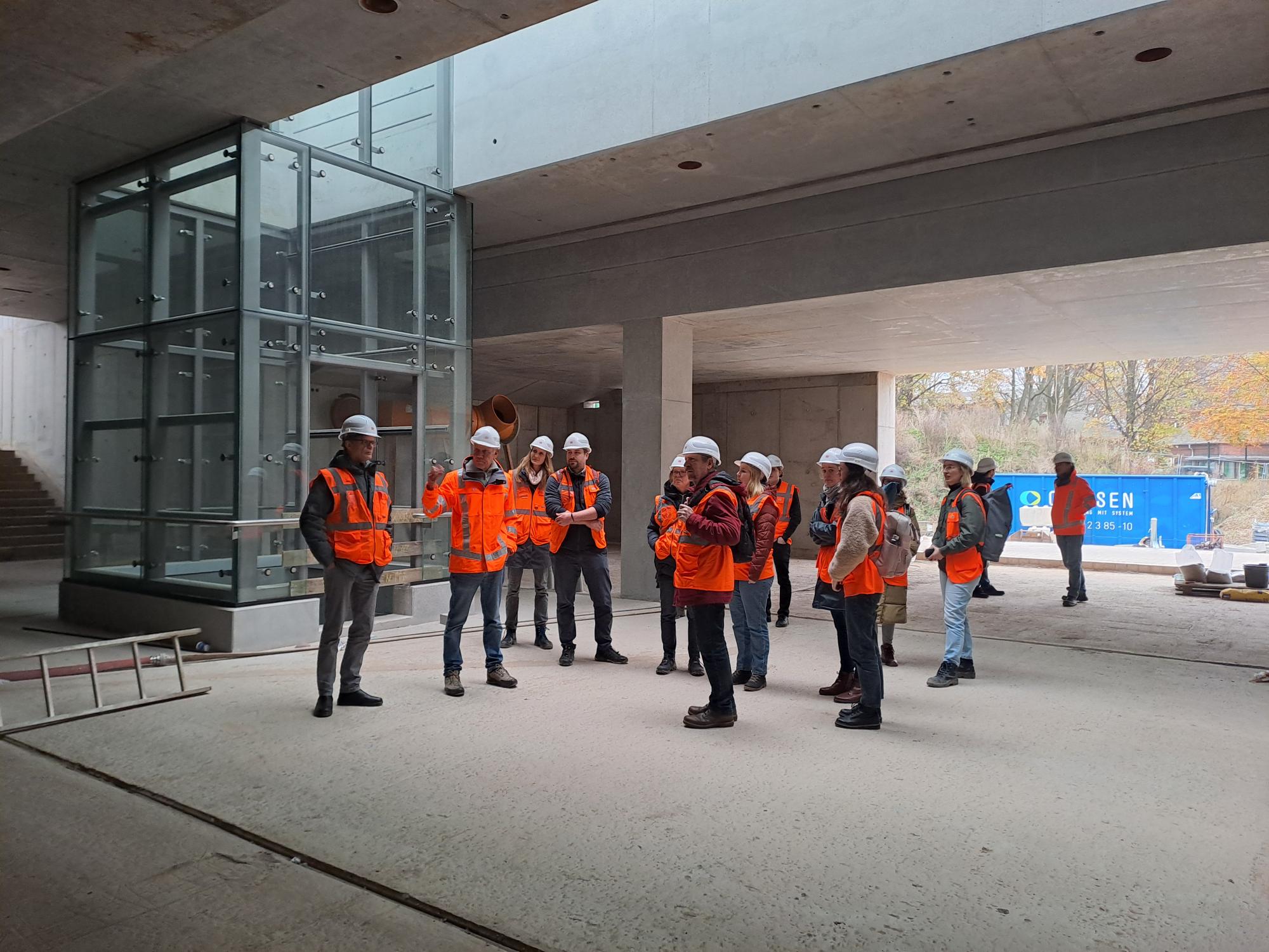 Eine Gruppe steht mit orangenen Warnwesten und Bauhelmen auf einer Baustelle.